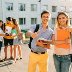 Happy two young students with note-books and backpacks smiling and showing thumb up Happy two young students with note-books and backpacks smiling and showing thumb up while standing on background of university and friends